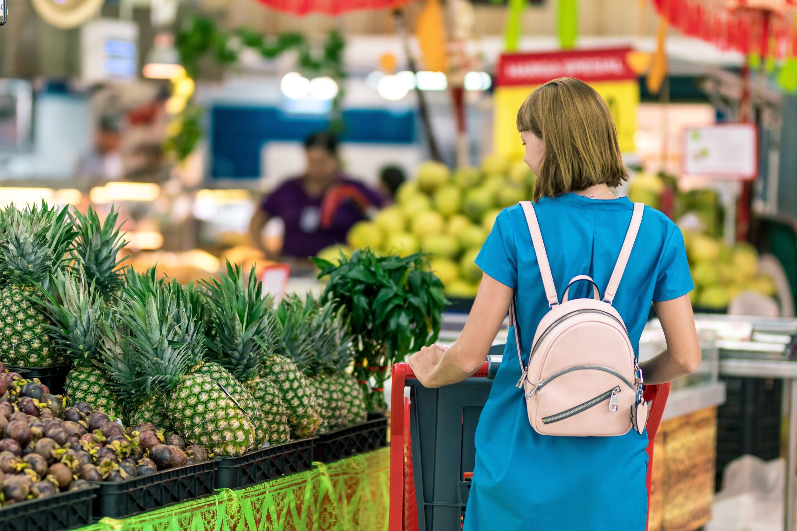 A woman in a blue dress and pink backpack explores a fruit market, standing before a display of pineapples and other fruits. The vibrant market, adorned with colorful banners, bustles around her as she navigates through it with the focus of an experienced supermarket liability attorney.