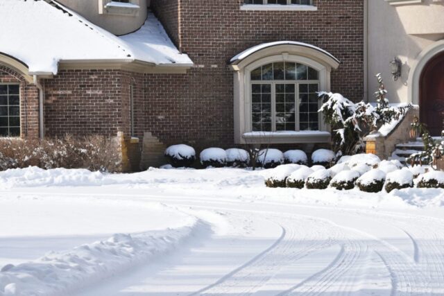 A brick house with snow-covered bushes, roof, and driveway evokes a crisp winter scene—reminiscent of those near ski resorts where ski accident lawsuit or ski resort injuries can sometimes occur. Tire tracks curve through the fresh snow in front.