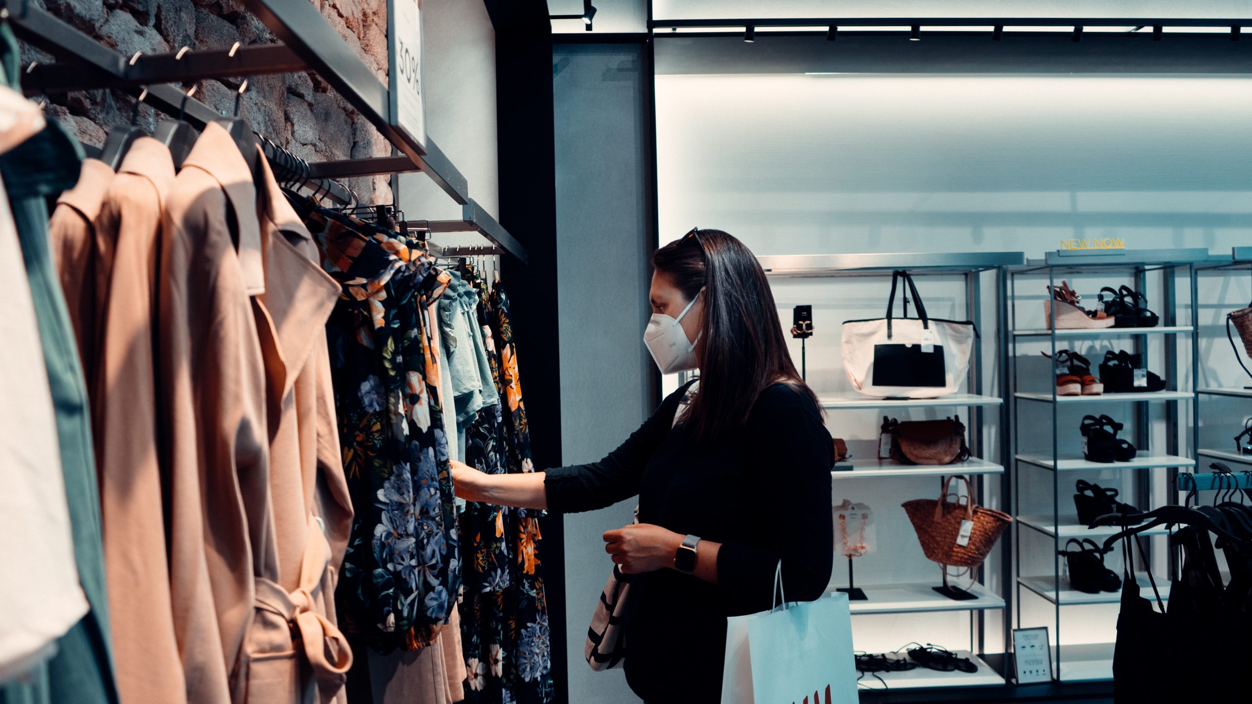 A woman wearing a mask is browsing clothing in a store. She is looking at a floral dress on a rack while holding a shopping bag. Shelves with handbags and baskets are visible in the background.