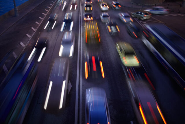 Blurred image of a busy urban street at night, with multiple cars and a bus in motion, creating streaks of light from headlights and taillights. Streetlights illuminate the scene, with a glimpse of water on the left.