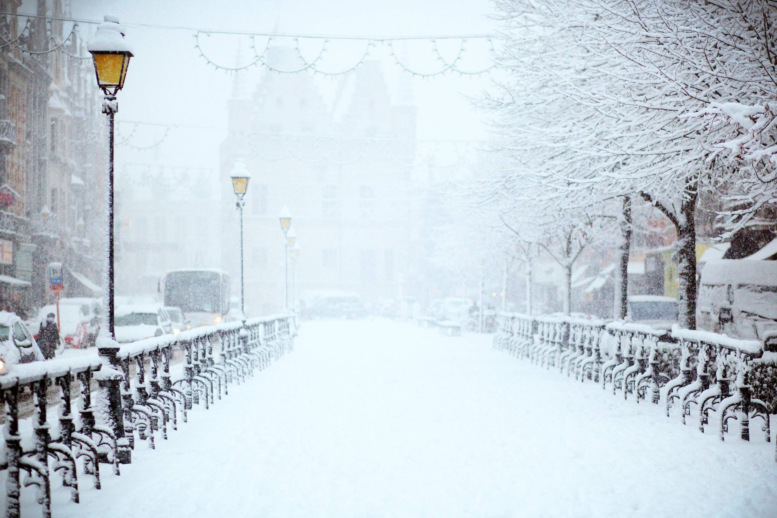 A snow-covered street in New Jersey features trees and lampposts lining both sides. Buildings in the background are partially obscured by falling snow. The scene is peaceful and wintry, despite the soft hum of an ice gritter ensuring no snow-and-ice accidents disturb this serene landscape.