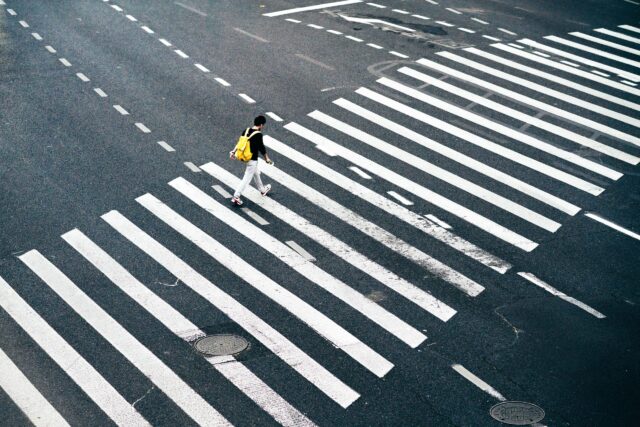 A person with a yellow backpack is walking across a large, multi-lane pedestrian crossing with white zebra stripes on an empty road, highlighting the importance of safe crossings to prevent pedestrian accidents. The scene is shot from an elevated viewpoint.