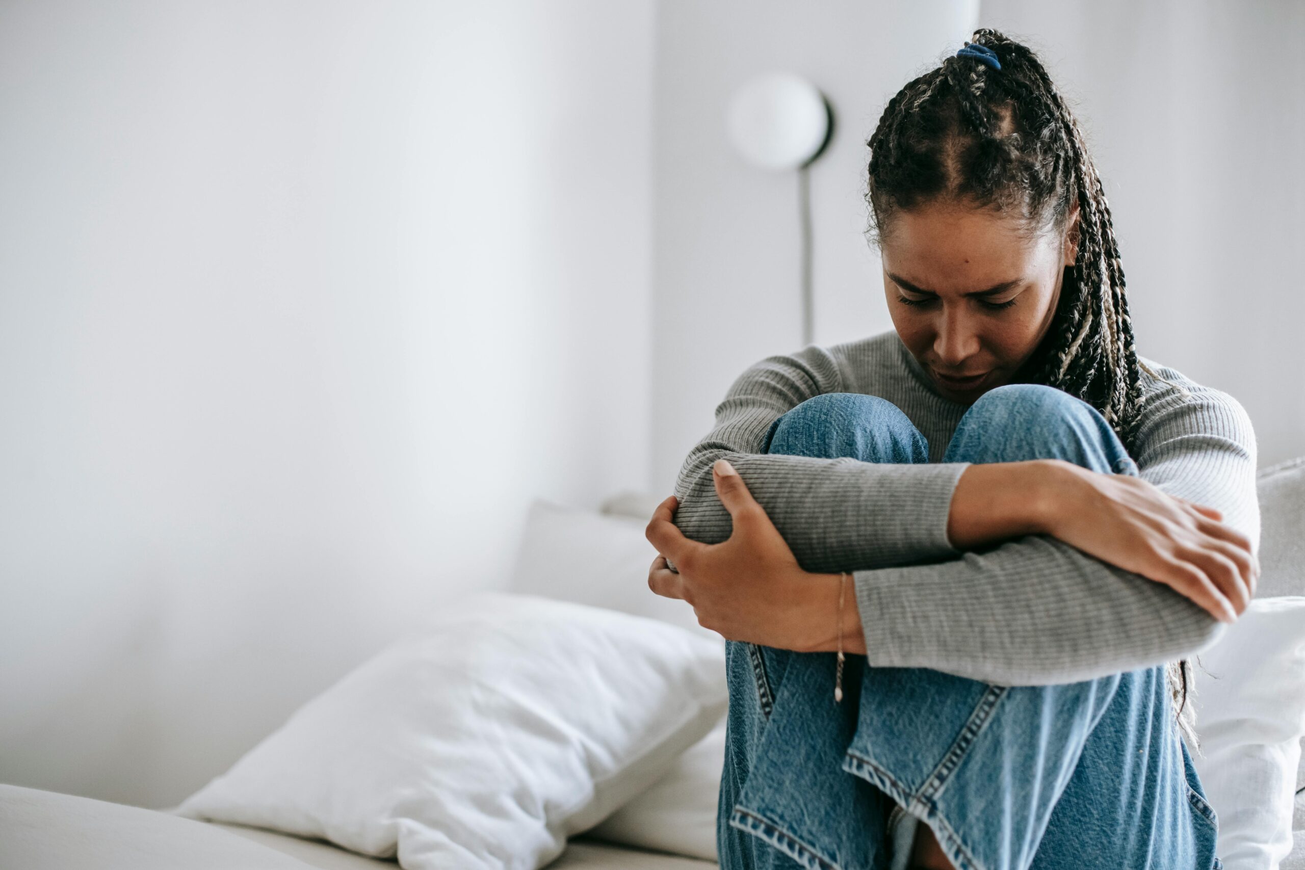 A woman sits on a bed hugging her knees, looking down with a sad expression. She is wearing a gray sweater and jeans, perhaps weighed down by concerns over New Jersey pain and suffering or challenges with insurance companies. White walls and pillows surround her.