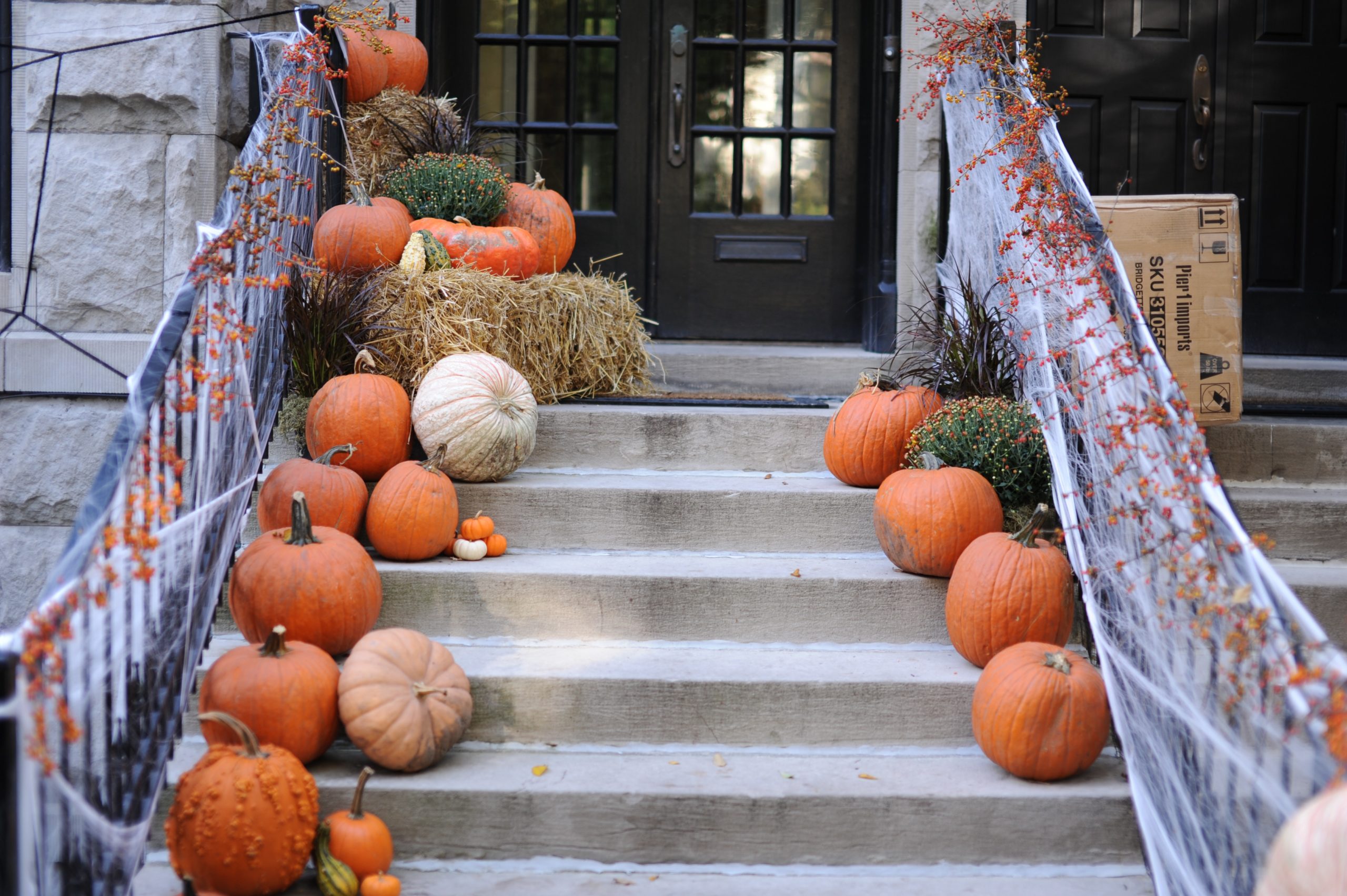 A staircase in New Jersey is decorated for autumn, with pumpkins of various sizes and colors lining both sides. Straw bales and dried branches enhance the seasonal theme, perfect for upcoming trick-or-treating. A cardboard box rests at the top of the stairs near the door.
