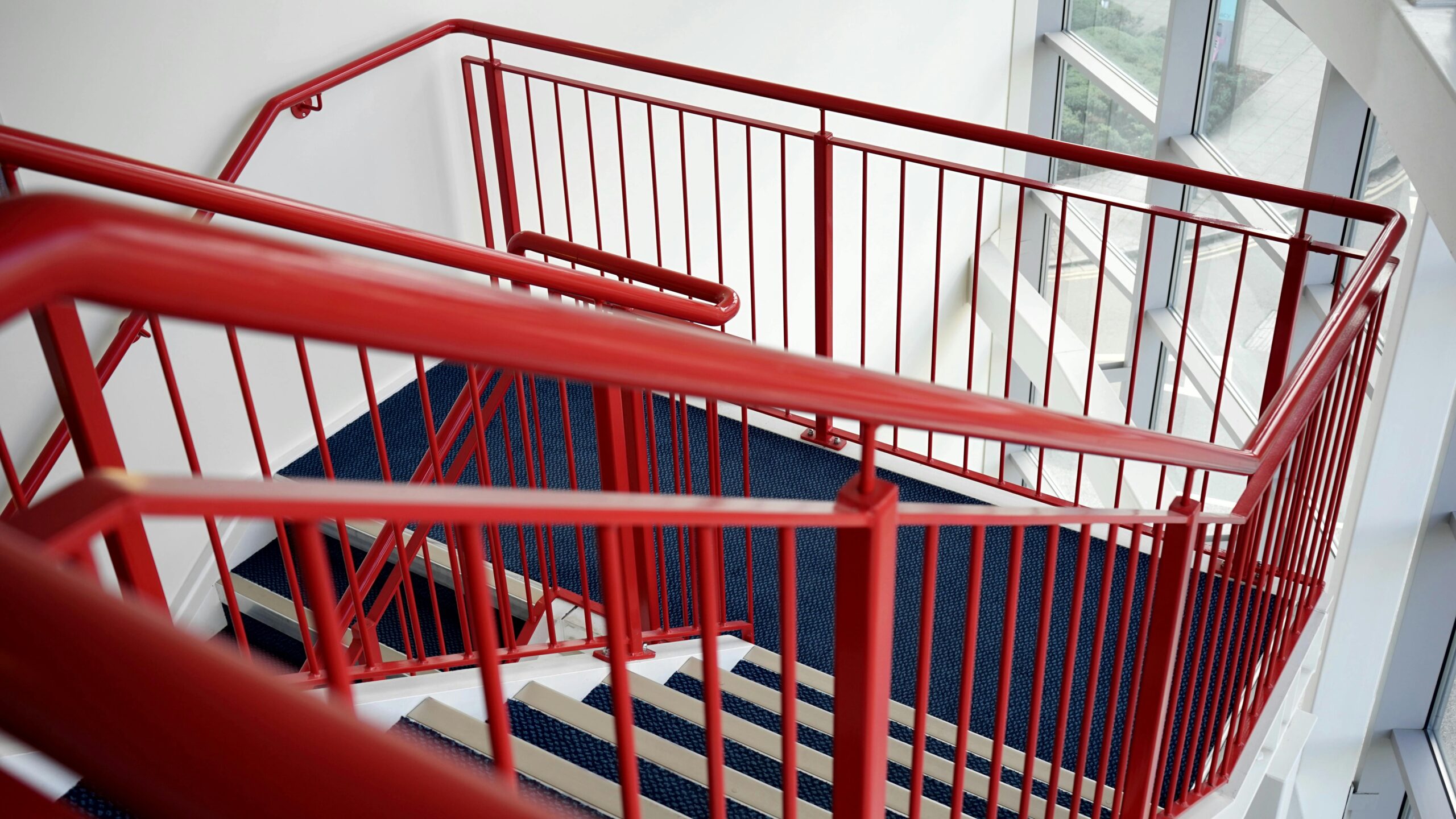 A staircase with vibrant red metal railings and dark blue carpeted steps, located indoors next to large windows letting in natural light—a reminder to stay cautious as accidents on broken stairs in New Jersey can lead to personal injury claims.
