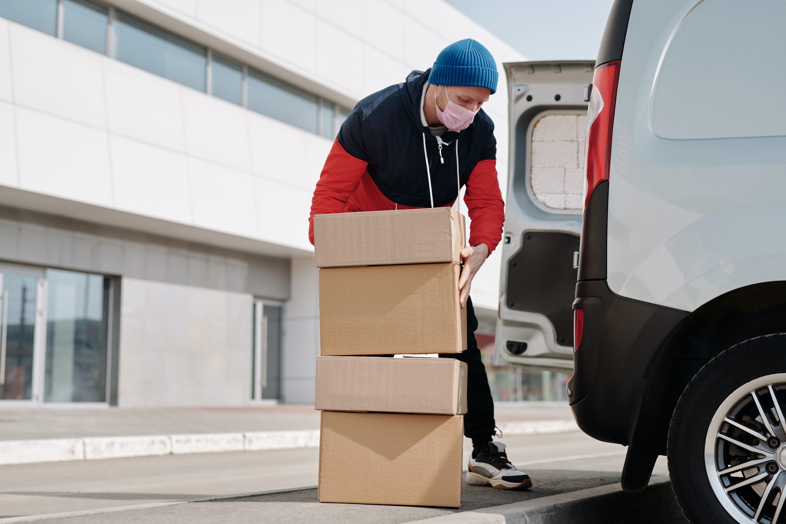 A person wearing a red and black hoodie, blue beanie, and face mask unloads cardboard boxes from the back of an Amazon delivery vehicle in an urban setting. They carefully place the boxes on the pavement, mindful of avoiding any potential delivery accidents.