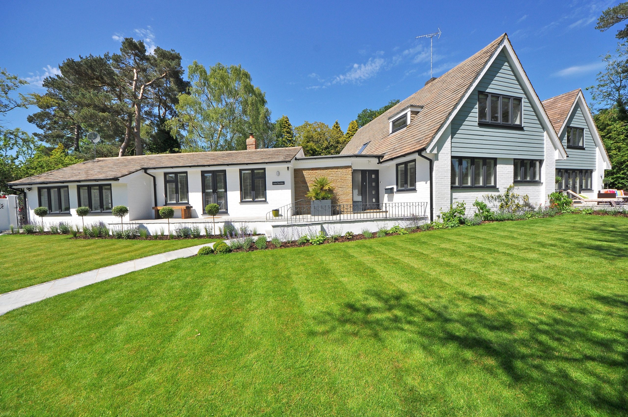 A modern, multi-story house with white walls and a gray gabled roof sits peacefully amidst a well-manicured lawn in New Jersey. The clear blue sky and tall trees enhance the property's serene setting, making it a comfortable retreat while emphasizing the importance of premises liability.