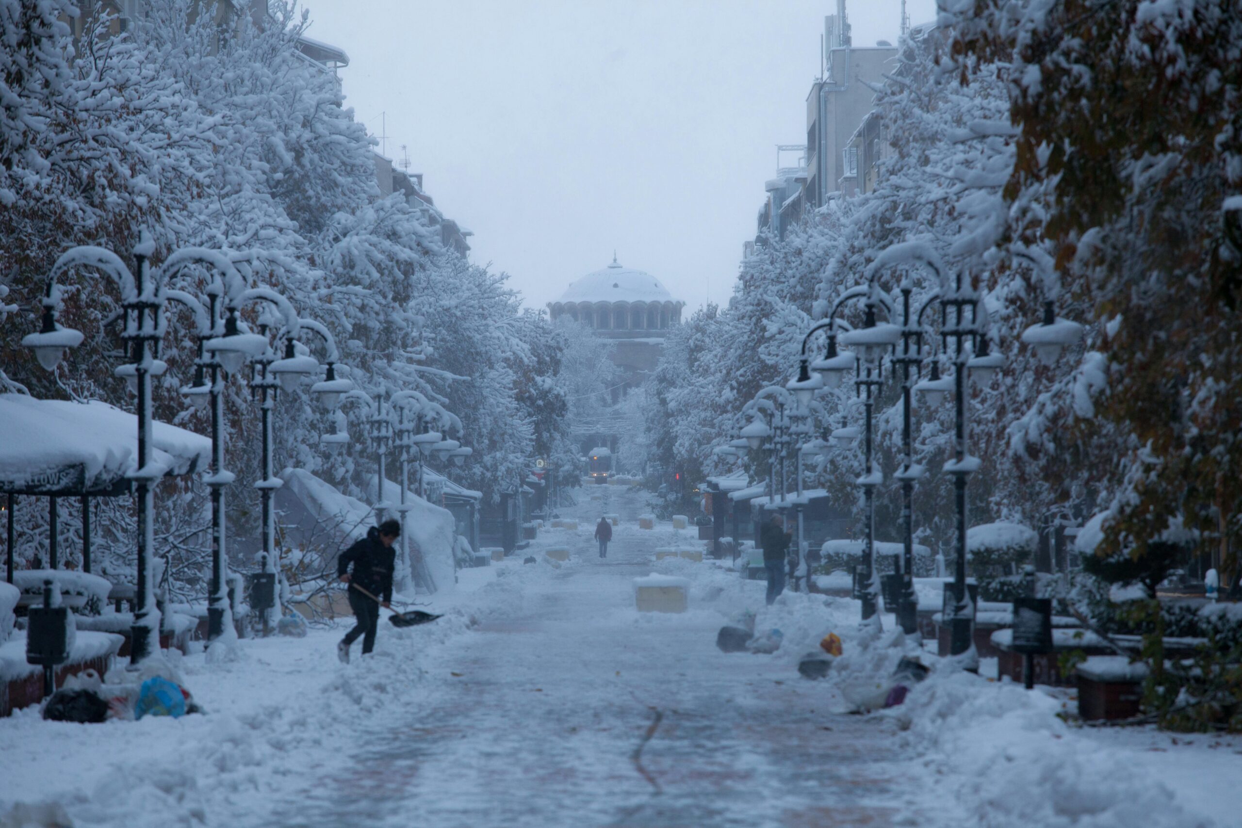 A snowy New Jersey city street lined with trees and lampposts is heavily covered in snow. A person handles snow removal on the left side, while others walk in the distance. A large domed building stands at the end of the street.
