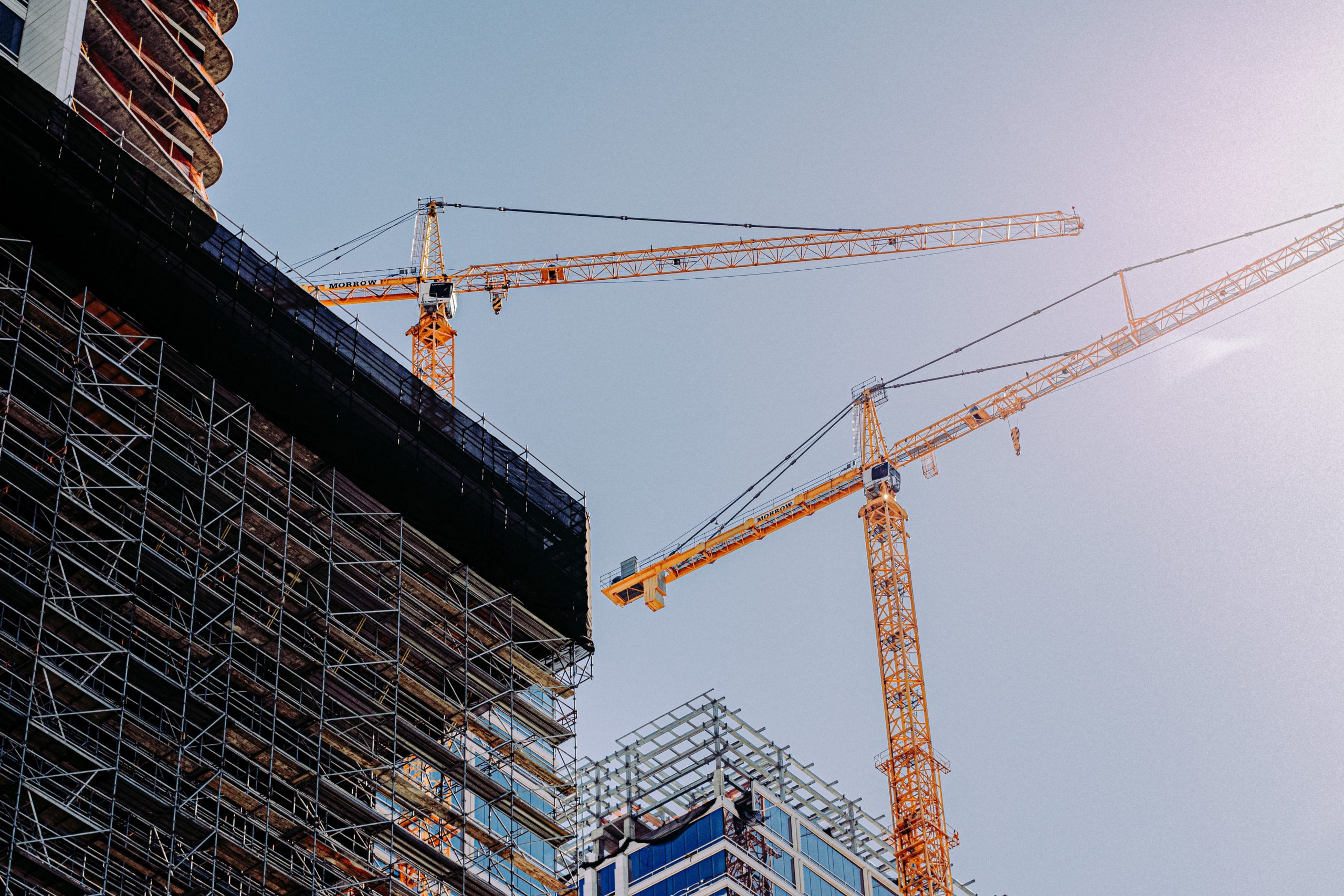 Two tall yellow construction cranes tower above buildings with visible scaffolding, embodying the bustling essence of a thriving construction site. They stand against a clear blue sky, where the sun gleams brightly overhead.