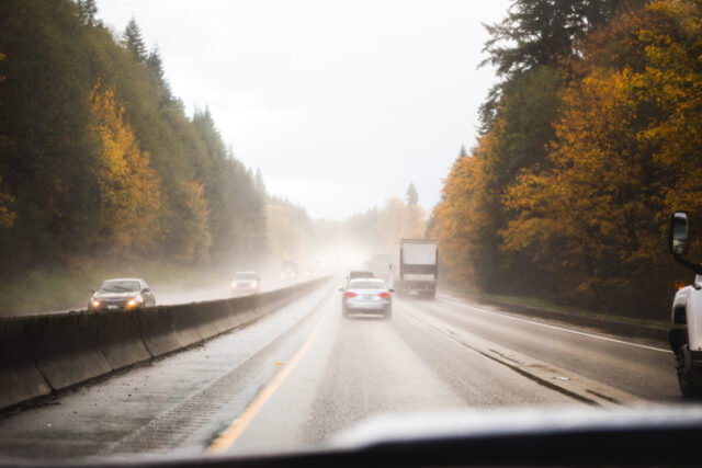 Cars and trucks drive on a wet highway surrounded by autumn trees in foggy weather; some vehicles have headlights on, and the concrete barrier helps reduce highway accidents.