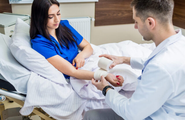 A doctor wraps a bandage around a woman’s forearm as she sits on a hospital bed, wearing blue scrubs and covered with a white blanket, after addressing concerns about potential doctor misdiagnosis.