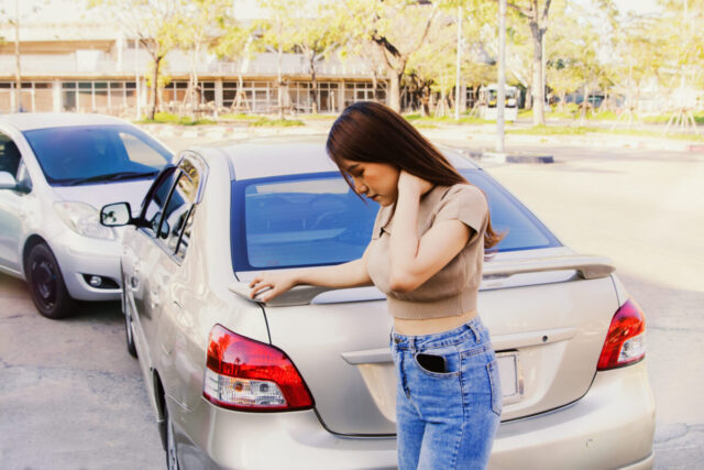 A young woman stands next to a silver car, looking concerned and holding her neck, while another car is parked closely behind hers; the scene highlights the importance of accident prevention in everyday driving.